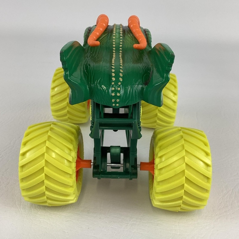 Pu&ograve; includere: Un giocattolo di monster truck verde con un design simile a una creatura sulla parte superiore, con corna arancioni e un corpo strutturato. Il camion ha quattro grandi pneumatici gialli con mozzi arancioni e un telaio verde.