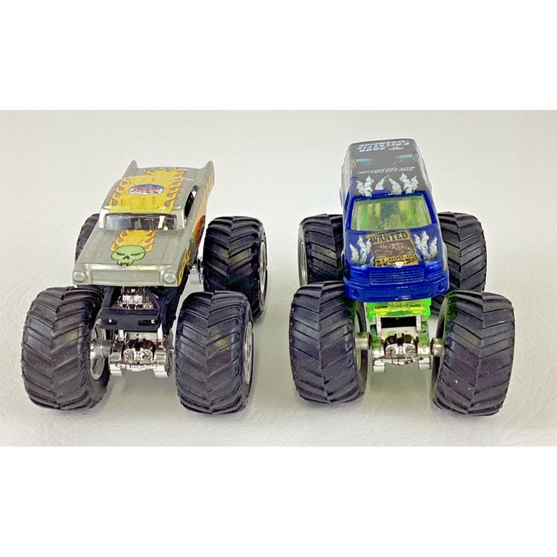 K&ouml;nnte beinhalten: Zwei Spielzeug-Monstertrucks. Einer ist silberfarben mit einem gelb-orangenen Design und einer Totenkopf-Grafik. Der andere ist blau und gr&uuml;n mit dem Wort "WANTED" auf der Windschutzscheibe. Beide haben gro&szlig;e schwarze Reifen.
