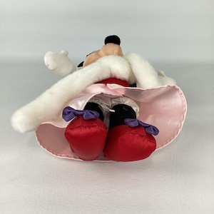 May include: A plush character toy featuring a black head with round ears, wearing a white faux fur-trimmed cape over a light pink satin skirt. The character has white gloves, black legs, red shoes with purple bows, and white ruffled bloomers.