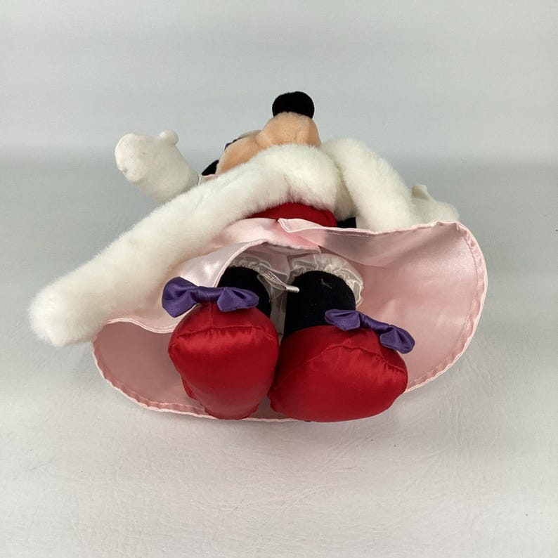 May include: A plush character toy featuring a black head with round ears, wearing a white faux fur-trimmed cape over a light pink satin skirt. The character has white gloves, black legs, red shoes with purple bows, and white ruffled bloomers.
