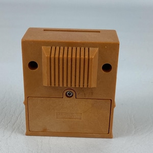 Op de afbeelding: Een kleine, bruin gekleurde, rechthoekige spaarpot met een gleuf aan de bovenkant voor het invoegen van munten. De achterkant heeft een textuurontwerp en een klein, rechthoekig batterijcompartiment. De bank is gemaakt van plastic en heeft twee ronde gaten aan de zijkanten.