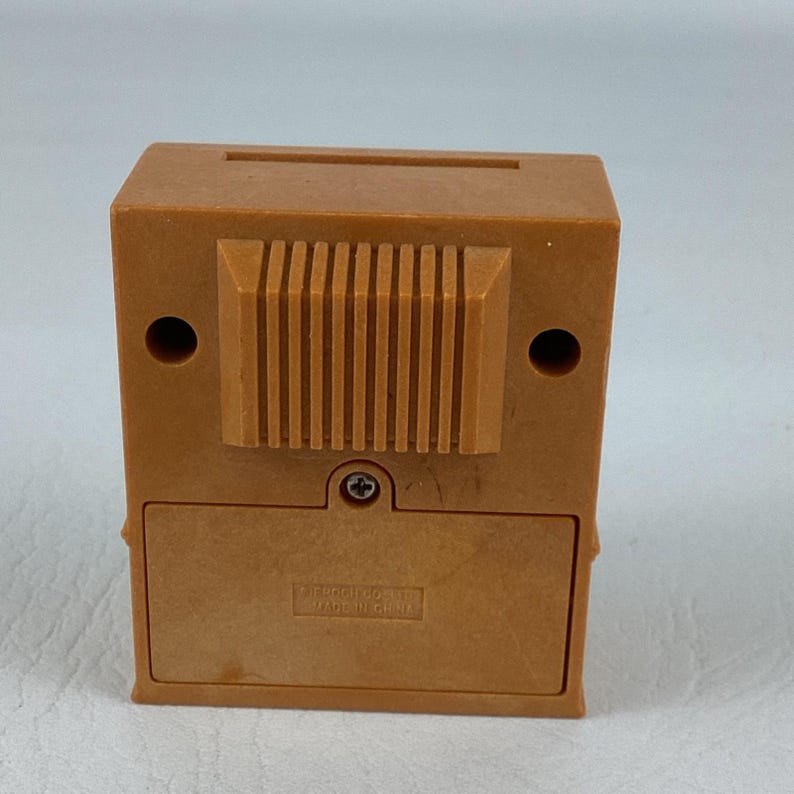 Op de afbeelding: Een kleine, bruin gekleurde, rechthoekige spaarpot met een gleuf aan de bovenkant voor het invoegen van munten. De achterkant heeft een textuurontwerp en een klein, rechthoekig batterijcompartiment. De bank is gemaakt van plastic en heeft twee ronde gaten aan de zijkanten.