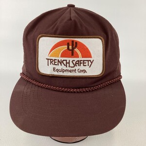 May include: A brown baseball cap featuring a white rectangular patch on the front. The patch displays an orange and yellow sunset graphic with a black cactus silhouette and the text TRENCH SAFETY Equipment Corp. in brown lettering.