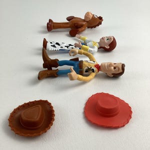 May include: A collection of Toy Story character figurines, including Woody, Jessie, and Bullseye, along with two cowboy hats. Woody is wearing a yellow shirt, blue jeans, and a sheriff's badge. Jessie has a white shirt with black cow spots. The hats are brown and red.