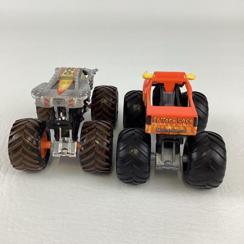 Puede incluir: Dos camiones monstruo de juguete. Uno es plateado con llamas naranjas y amarillas, el otro es naranja con el texto "El Toro Loco" y "MonsterJam.com". Ambos tienen neum&aacute;ticos negros grandes.
