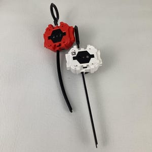 May include: Two plastic toy launchers, one red and one white, with black pull cords and loop handles. The launchers have a geometric design with black accents and a QR code.