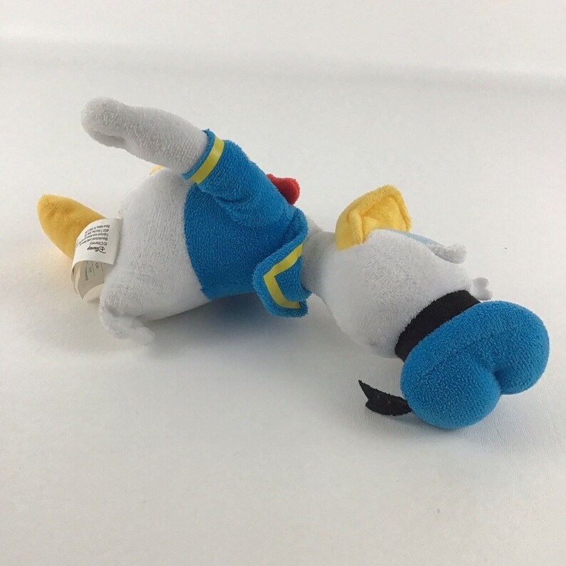 May include: A white and yellow plush toy of Donald Duck wearing a blue sailor shirt with yellow trim and a black sailor hat. The toy is lying on its side with its head turned to the right.