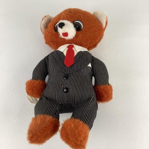 Pode incluir: Um urso de pel&uacute;cia vintage vestindo um terno listrado preto e branco com uma gravata vermelha. O urso tem pelo laranja na cabe&ccedil;a, patas e p&eacute;s. Possui gola branca e um pequeno len&ccedil;o de bolso branco.