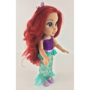May include: A red-haired doll wearing a purple and green mermaid costume with a crown. The doll has blue eyes and a smiling face.