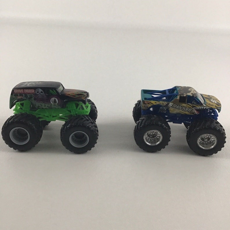 May include: Two monster truck toys, one black and green with "Grave Digger" written on the side, and the other blue, yellow, and green with "Shocker" written on the side.