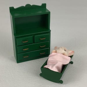 May include: Miniature green wooden dresser with three drawers and a top shelf, next to a green cradle with a pink blanket and a small stuffed animal inside. The furniture is painted in a dark green color.