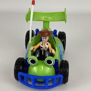 May include: A green and blue toy race car featuring the character Woody from Toy Story. The car has large black tires, a blue front bumper, and a green spoiler. Woody is wearing a cowboy hat and a yellow shirt.