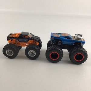 May include: Two monster trucks, one orange and black with "Predator" written on the side, and the other blue with orange flames, both with large black tires.