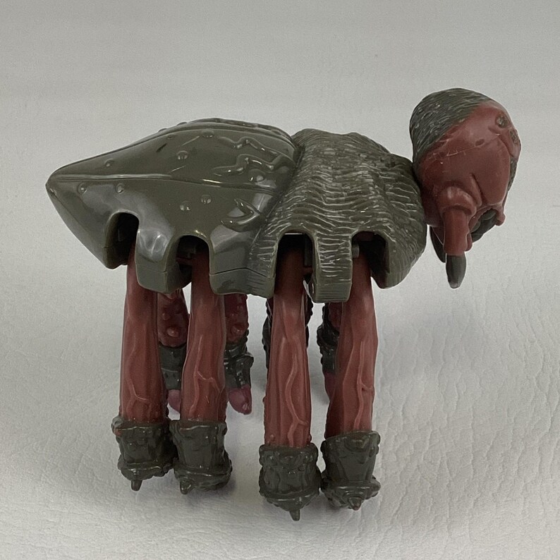 K&ouml;nnte beinhalten: Eine mehrbeinige Kreaturen-Actionfigur mit einem glatten, olivgr&uuml;nen Panzer, einem texturierten, gerippten Mittelteil und einem rotbraunen Kopf. Die Figur steht auf mehreren langen, rotbraunen, aderartigen Beinen mit dunkel olivgr&uuml;nen, gepanzerten F&uuml;&szlig;en.