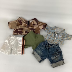 Puede incluir: Un conjunto de ropa infantil, que incluye una camisa de camuflaje, una camisa estampada azul, pantalones cortos blancos con una raya naranja, pantalones cortos verde oliva y pantalones cortos de mezclilla con dobladillos.