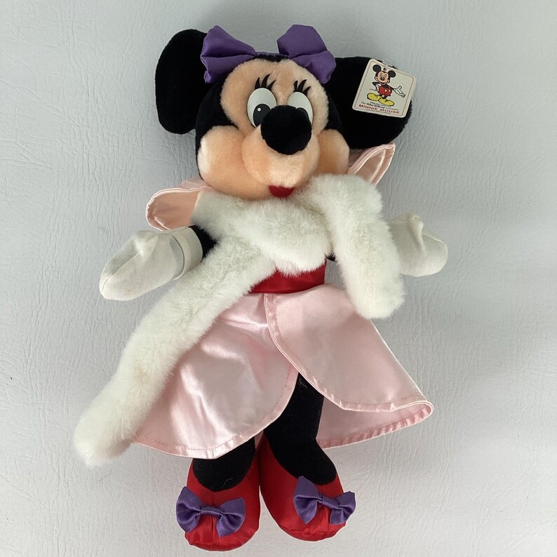May include: A plush Minnie Mouse doll featuring a black head with a purple bow, wearing a light pink satin dress with a white faux fur stole. The doll has white gloves and red shoes with small purple bows, and includes a Disney brand tag attached.