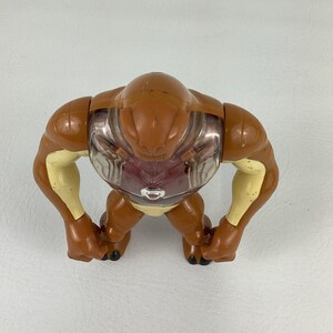 May include: A brown and cream-colored plastic action figure with a smooth, rounded head and muscular arms. The torso features a clear, transparent chest plate revealing a dark red interior, with a small silver circular emblem centered on the chest.