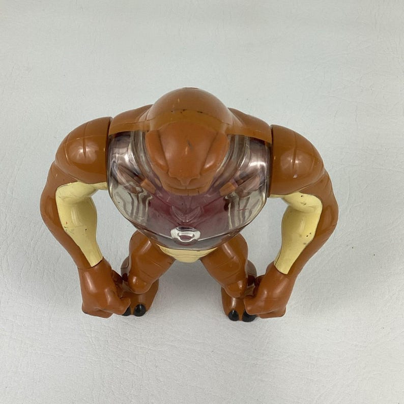 May include: A brown and cream-colored plastic action figure with a smooth, rounded head and muscular arms. The torso features a clear, transparent chest plate revealing a dark red interior, with a small silver circular emblem centered on the chest.