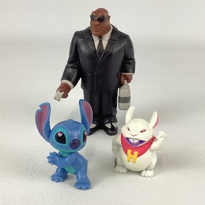 May include: A group of three figurines: a black-suited character with sunglasses, a blue alien-like creature, and a white rabbit-like figure with a red scarf and a yellow "H" on it. The figures are on a white surface.