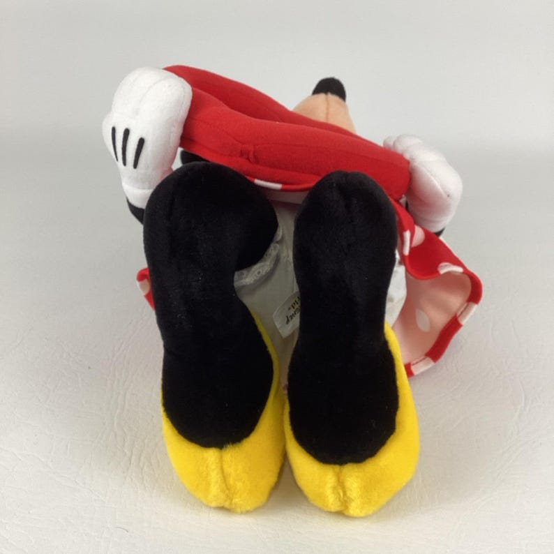 Pu&ograve; includere: Un peluche con un vestito rosso a pois bianchi, piedi neri con suole gialle, guanti bianchi e un naso nero. Il giocattolo &egrave; sdraiato sulla schiena, con le braccia e le gambe in aria.