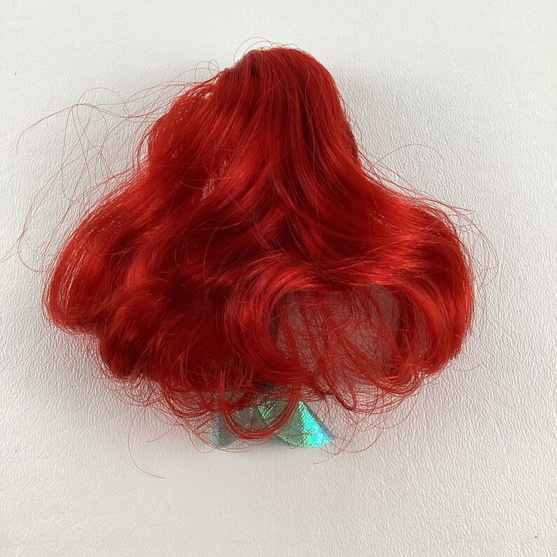 Puede incluir: Una peluca roja de mu&ntilde;eca con cabello largo y suelto. La peluca es de un tono rojo vibrante y parece estar hecha de fibras sint&eacute;ticas. La peluca est&aacute; peinada con ondas y rizos sueltos. La peluca es probablemente para una mu&ntilde;eca o un disfraz.