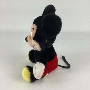 May include: A plush Mickey Mouse toy, featuring black fur, a red shorts, and yellow shoes. The toy has a white face and hands, with a black tail. The classic character is sitting down, with a side profile view.