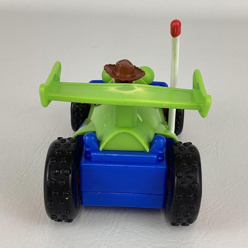 May include: A toy race car with a green body and spoiler, blue chassis, and black tires. A small brown hat sits atop a green figure in the driver's seat. A white antenna with a red tip extends from the car.
