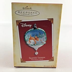 Peut inclure: Une boule de Noël Hallmark Keepsake représentant une boule de Noël blanche, bleue et verte avec une scène de Bambi et de Panpan faisant du patin à glace. La boule est dans sa boîte d'origine, qui est rouge et blanche avec les mots "Disney" et "Leçon de patinage Bambi de Walt Disney" imprimés dessus.