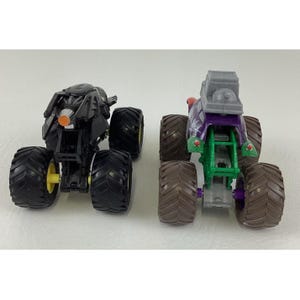 May include: Two toy monster trucks. One is black with yellow accents, the other is purple and green with brown tires. Both have large, black, treaded tires. The trucks are on a white surface.