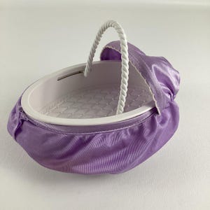 May include: A purple and white baby doll bassinet with a white rope handle. The bassinet has a quilted lining and a soft fabric exterior.