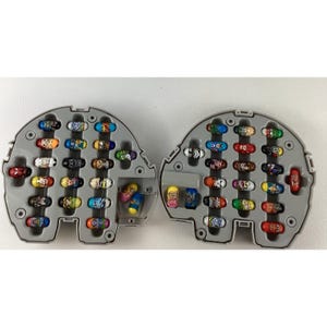 May include: Two grey plastic storage cases, each featuring multiple moulded slots containing small, oval-shaped collectible figures with various colourful character designs. The figures are arranged in rows within the open, semi-circular containers.