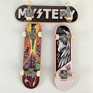 May include: Three finger skateboards with grey trucks and white wheels. The top board is black with the white text MYSTERY. The bottom left board features a colourful graphic with yellow, red, and green tones. The bottom right board has a black and white eagle graphic and the text birdhouse.