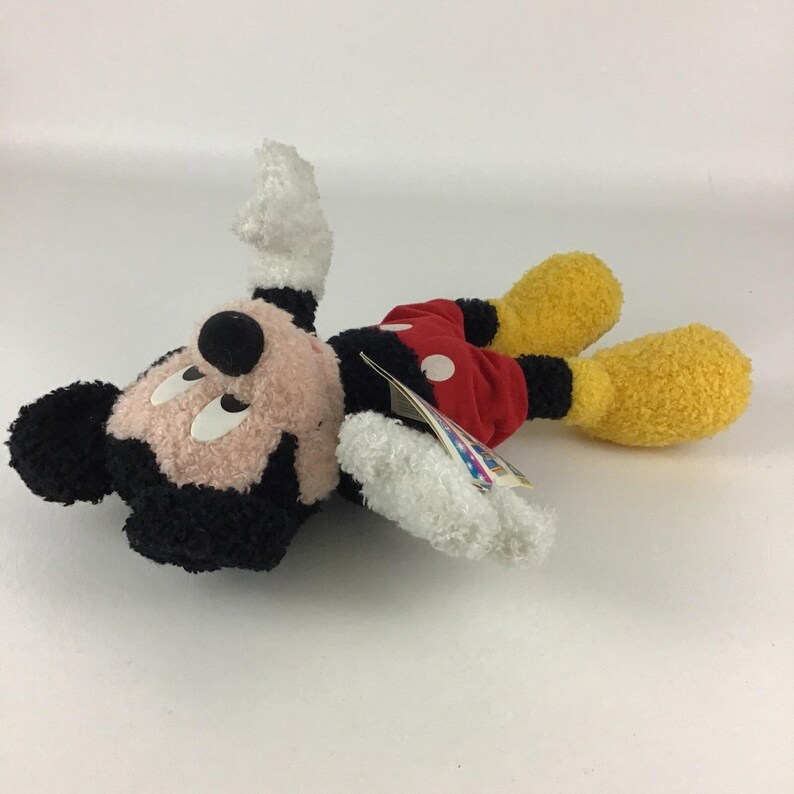 May include: A plush Mickey Mouse toy, lying on its side. The toy features a black head and ears, a pink face, and white gloves. It wears red shorts with white buttons and yellow feet. A tag is attached.