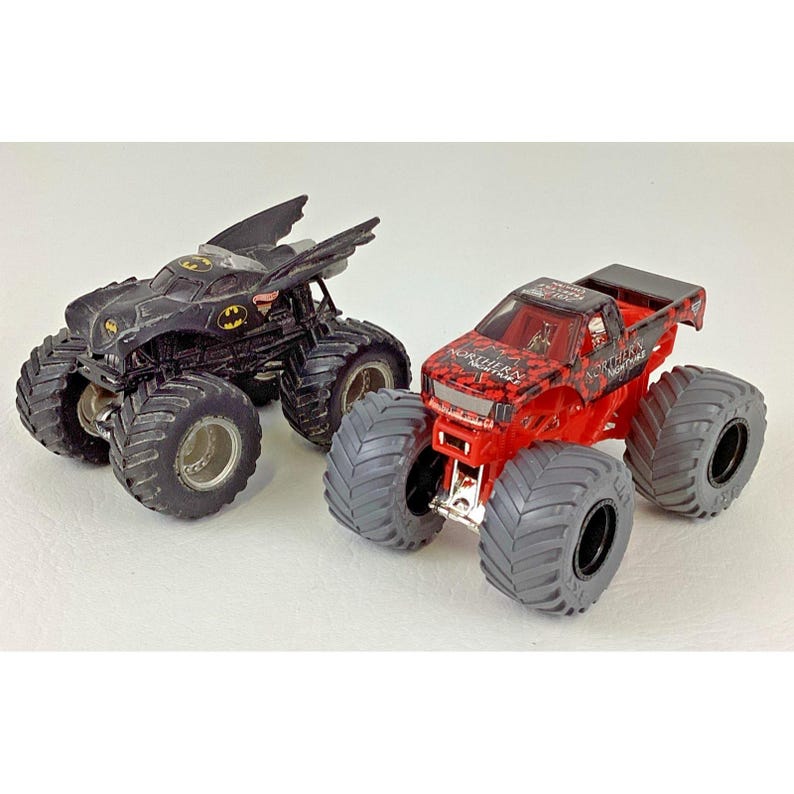 May include: Two toy monster trucks. One is black with a Batman design, featuring large tires and a bat-shaped wing design. The other is red with black accents and text on the side, also with large tires.
