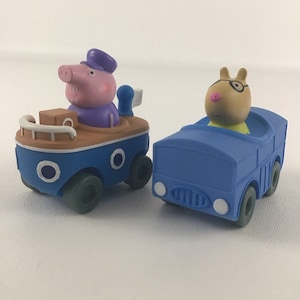 May include: Two toy vehicles featuring characters from the children's television show Peppa Pig. A blue boat with a brown deck and a brown pig wearing a blue hat driving it. A blue bus with a brown pig wearing glasses driving it.
