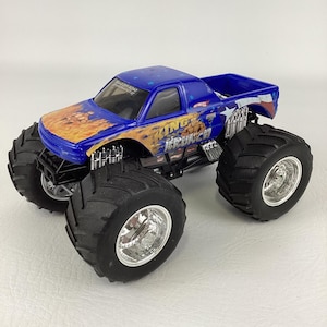 Op de afbeelding: Een blauw en oranje Hot Wheels King Krunch monstertruck speelgoed. De truck heeft grote zwarte banden met zilveren velgen en de woorden "King Krunch" op de zijkant. De truck heeft een blauwe cabine en laadbak.