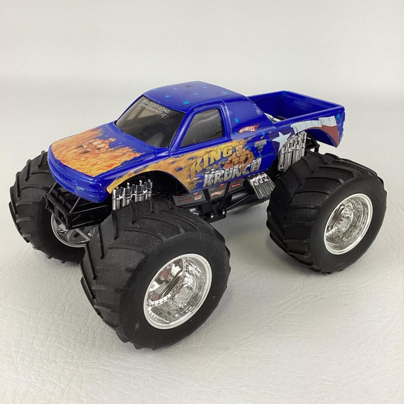 Op de afbeelding: Een blauw en oranje Hot Wheels King Krunch monstertruck speelgoed. De truck heeft grote zwarte banden met zilveren velgen en de woorden "King Krunch" op de zijkant. De truck heeft een blauwe cabine en laadbak.