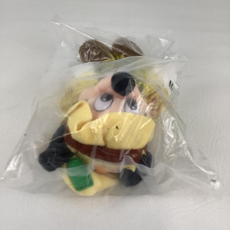 K&ouml;nnte beinhalten: Ein Pl&uuml;schtier mit gelbem Hut, braunem Haar und gro&szlig;en wei&szlig;en Augen mit schwarzen Pupillen, verpackt in einer durchsichtigen Plastikt&uuml;te. Die Figur hat eine schwarze Nase, einen gelben Kragen und ein gr&uuml;nes rechteckiges Element im unteren Bereich.