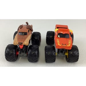May include: Two toy monster trucks. One is brown and tan, shaped like a dog with a red tongue. The other is orange with yellow horns and a flame design. Both have large black tyres and are on a white surface.