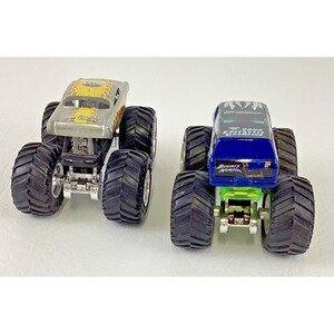 K&ouml;nnte beinhalten: Zwei Spielzeug-Monstertrucks. Einer ist silberfarben mit gelben und orangen Flammen und dem Text "Racing". Der andere ist blau mit dem Text "Bounty Hunter" und "2000 Freestyle Champion". Beide haben gro&szlig;e schwarze Reifen.