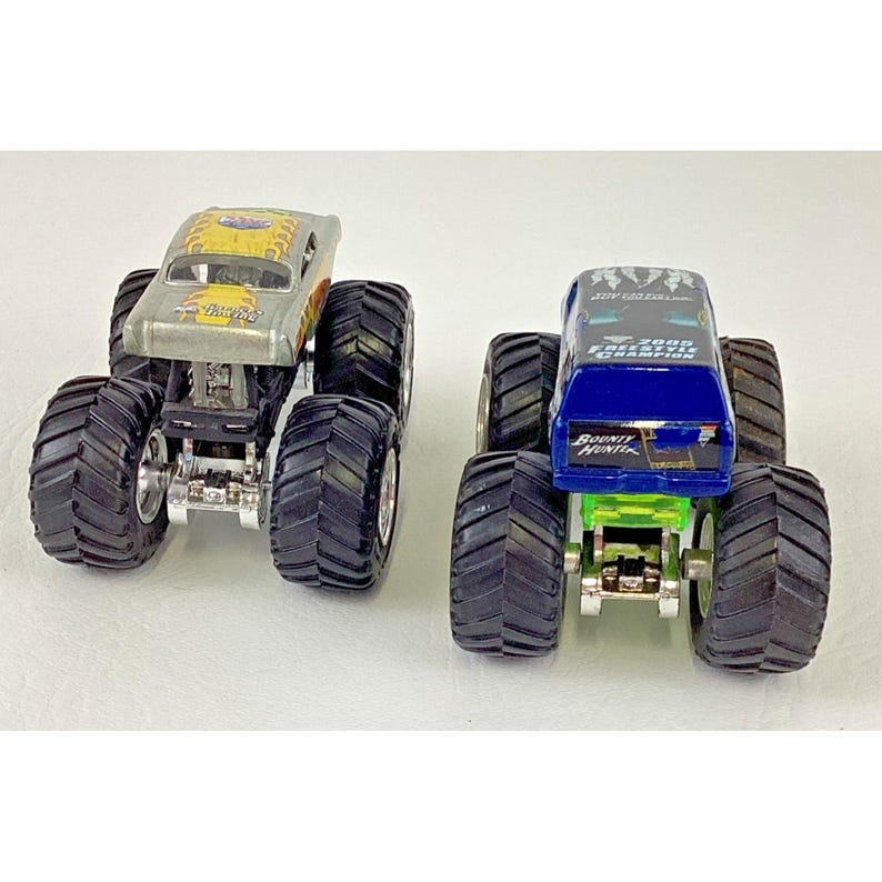 K&ouml;nnte beinhalten: Zwei Spielzeug-Monstertrucks. Einer ist silberfarben mit gelben und orangen Flammen und dem Text "Racing". Der andere ist blau mit dem Text "Bounty Hunter" und "2000 Freestyle Champion". Beide haben gro&szlig;e schwarze Reifen.