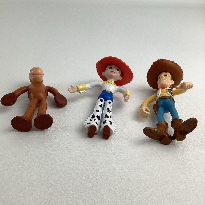 May include: Three plastic toy figures from the animated film Toy Story. The figures include a brown horse, a cowgirl with a red hat and cow print pants, and a cowboy with a brown hat and boots. The toys are on a white surface.