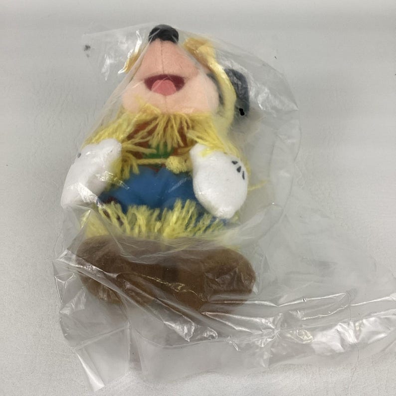 K&ouml;nnte beinhalten: Ein Pl&uuml;schtier mit hellem Gesicht, gelben Wollhaaren, wei&szlig;en Handschuhen, blauer Hose und braunen Schuhen, verpackt in einer durchsichtigen Plastikt&uuml;te. Die Figur hat einen weit ge&ouml;ffneten Mund und ist durch die Verpackung deutlich zu erkennen.