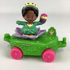 May include: A Little People toy featuring a princess figure in a green dress and a yellow crown, holding a cupcake. The princess sits in a purple flower on a green carriage with pink wheels. The carriage has a frog emblem.