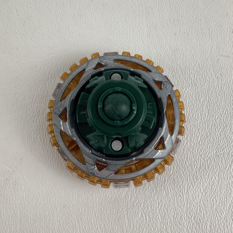 Puede incluir: Una peonza de juguete verde, plateada y dorada. El centro es de color verde oscuro, rodeado por un anillo plateado con bordes dorados en forma de dientes. El juguete est&aacute; dise&ntilde;ado para girar y jugar.