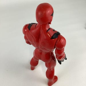 Puede incluir: Figura de acci&oacute;n roja con detalles negros en hombros y brazos. La figura se ve desde atr&aacute;s, mostrando su musculatura. El juguete est&aacute; hecho de un material pl&aacute;stico brillante.