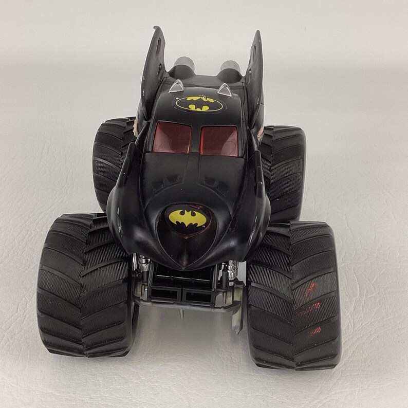 May include: Black Batman monster truck toy with large, black tires. The truck features the Batman logo in yellow on the front and top. The windows are red. The truck has large, bat-like fins on top.