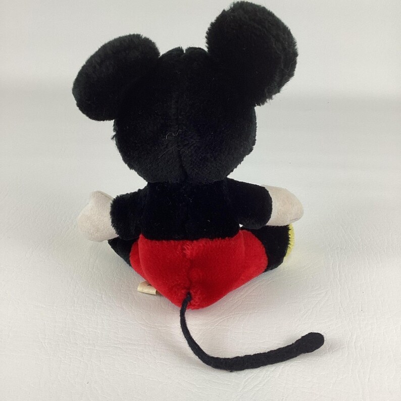 May include: A plush Mickey Mouse toy, viewed from the back, with black fur, large round ears, and a long tail. The toy wears red shorts, white gloves, and has a yellow patch on the bottom.