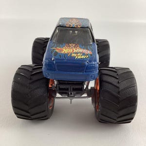 Puede incluir: Un juguete de cami&oacute;n monstruo Hot Wheels azul con neum&aacute;ticos negros grandes y llantas naranjas. El cami&oacute;n tiene un dise&ntilde;o de llama en el techo y las palabras "Hot Wheels Beat That!" en la parte delantera.