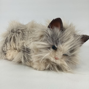 Op de afbeelding: Een pluche kat met een pluizige, meerkleurige grijze en crèmekleurige vacht. De kat heeft blauwe ogen, een roze neus en lange witte snorharen. De oren zijn donkerbruin.